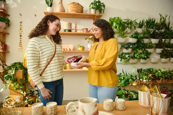 Two women in a plant store transacting with a mobile POS device. One woman pays. The other woman holds a white card reader.